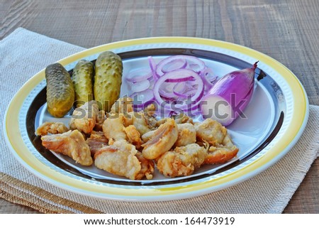 Plate of fried pieces of pork scratchings with red torpedo onions and pickles on wooden table.