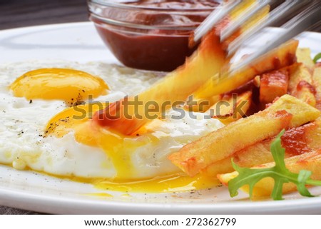 Fried eggs with french fries and ketchup. Dipping potato chips in fried egg yolk (motion blur).