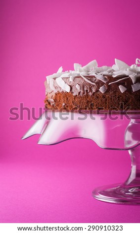 Chocolate cake with coconut flakes on the glass cake plate with purple background closeup vertical