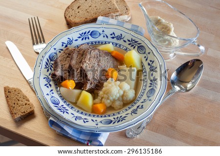 Boiled beef Tafelspitz soup broth with potato, carrot, cauliflower, kohlrabi, bred and horseradish sauce in glass sauce boat with cutlery and napkin on the wood table. Germany traditional cuisine