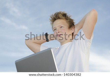 Resting teenager with laptop holding hands behind his head against blue sky (high key)