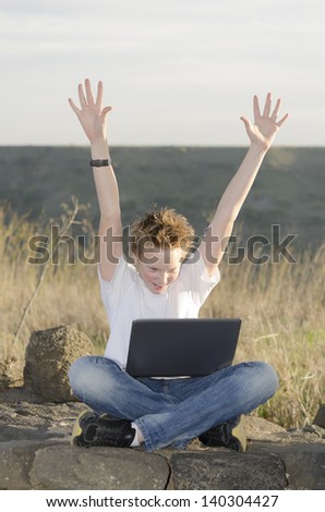 Shaggy teenager with laptop joyously throws his hands up in nature