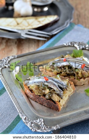 Bruschetta with marinated anchovies, baked eggplant and capers on a beautiful metal plate.