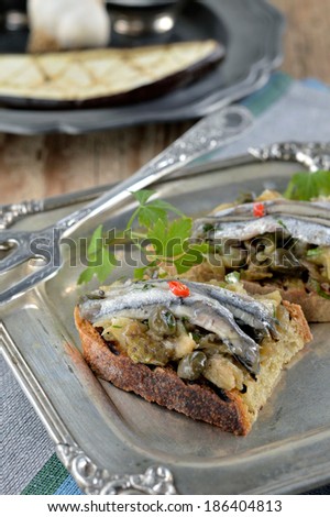 Bruschetta with marinated anchovies, baked eggplant and capers on a beautiful metal plate. Closeup.