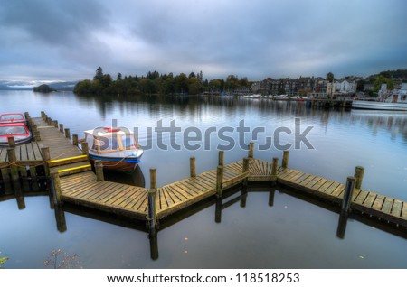 Landscape image of a wooden jetty on Lake Windermere in Lake District, a National Park in England, during Autumn with grey sky and colourful boats next to jetty