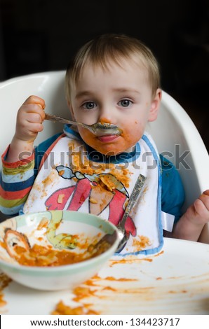 A very messy cute eight month old child trying to eat orange colored pasta
