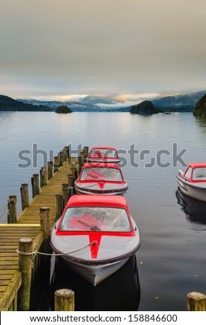 Landscape image of a wooden jetty on Lake Windermere in Lake District, a National Park in England, during Autumn with grey sky and colourful red boats next to jetty