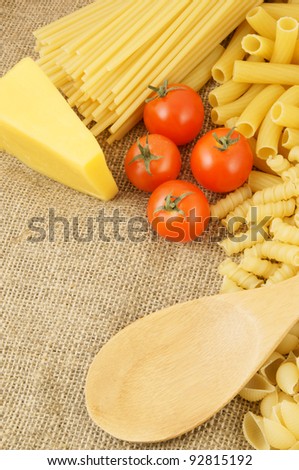 Raw macaroni with tomatoes, spoon and cheese on burlap background