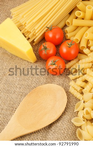 Raw macaroni with tomatoes, spoon and cheese on burlap background