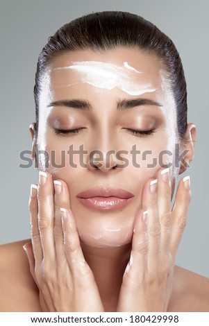 Beautiful healthy young woman with eyes closed while applying moisturizer to her clean face using gentle soft touches, close-up portrait on grey. Perfect skin. Skincare concept.