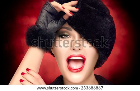 Winter Beauty Fashion. Close up portrait of pretty young woman with open mouth facial expression, wearing trendy fur hat and mittens. Gestures and grimaces. High fashion portrait on red background.