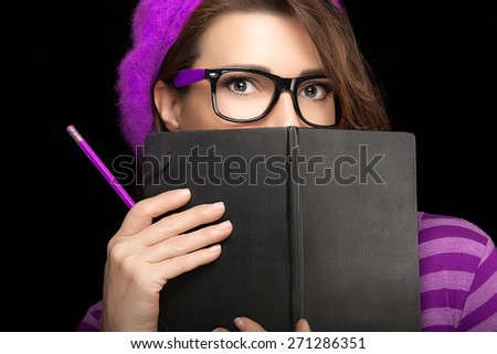 Close up Beauty Stylish College Girl with Expressive Eyes in Trendy Eyeglasses Covering her Face with Black Notebook While Holding a Pencil. Isolated on Black Background