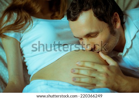 Image of happy future dad kiss the belly of his cheerful caucasian pregnant wife lying on bed at home.