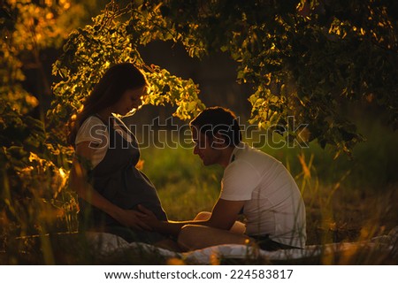 Image of happy future dad touching the belly of his beautiful caucasian pregnant wife relaxing on green grass in the evening. Young family is resting in the garden or park into the sunset.