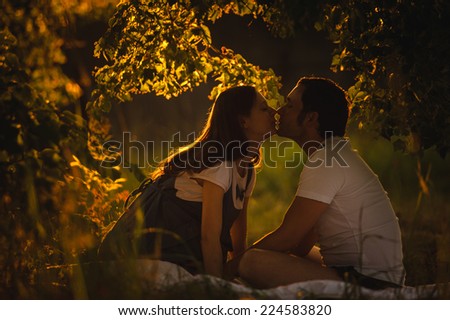Image of happy future dad kiss the belly of his beautiful caucasian pregnant wife relaxing on green grass in the evening. Young family is resting in the garden or park into the sunset.