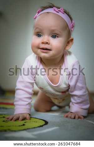 Adorable baby girl crawls on all fours on floor at home. Smiling infant of 6 months playing on ground and looking at camera. Portrait of female child. Vertical, front view. Funny kid goes forward
