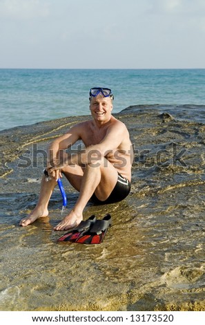 A happy laughing 44 year old man is sitting on a boulder at the beach during his vacation.