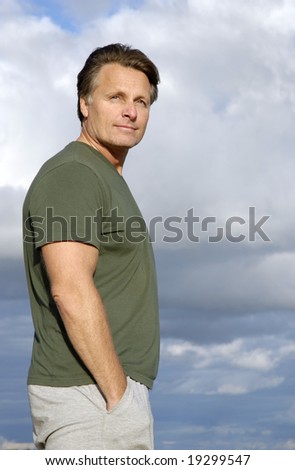 A handsome fit forties man is standing and watching with a cloudy sky behind him