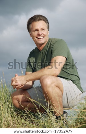 a happy handsome fit forties man sitting on the beach and enjoying his day out.Dark storm clouds are approaching from behind him.