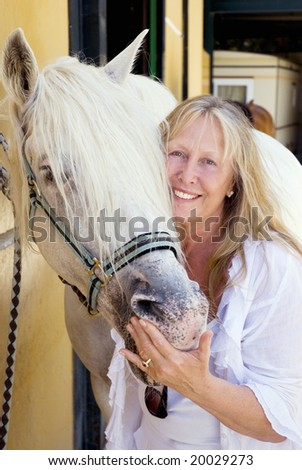 A beautiful mature woman in her sixties stands next to a white andalusian stallion with a happy smile on her face