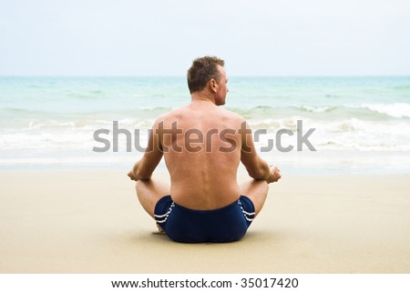 A mature man sitting cross legged on a sandy beach and meditating.