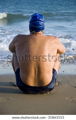 A male swimmer with a wide muscular back is sitting on the beach looking at the sea.