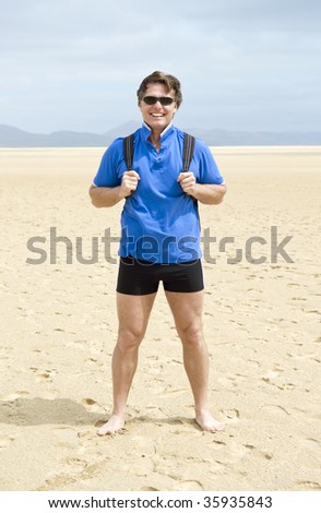 A colour portrait of a mature male backpacker standing on large empty beach.