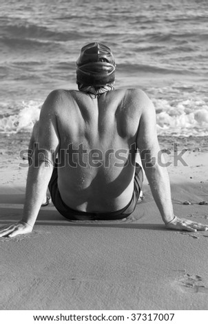 A black a white photo of a male swimmer sitting on the beach.