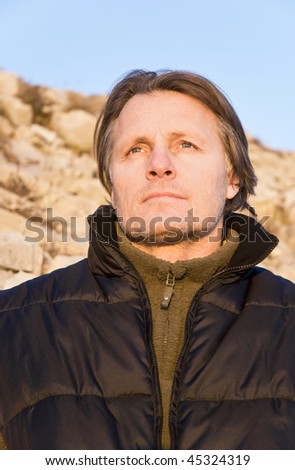 A pensive looking forties male with stubble looks out at the setting sun after a day spent walking in the mountains.