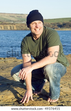 A colour portrait photo of a happy smiling forties man wearing a hat crouching down next to th the sea.