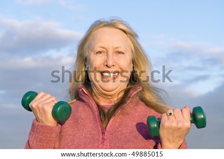 A colour portrait photo of a happy older woman exercising with dumbells on the beach.