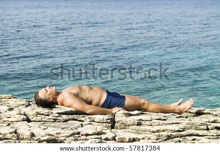 A color landscape photo of a caucasian man sunbathing on rocks next to the blue sea.