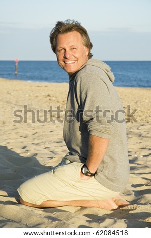 A color portrait photo of a happy smiling man in his forties kneeling down on the beach.