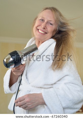 A color portrait photo of a beautiful blonde older woman smiling as she blow dries her hair.