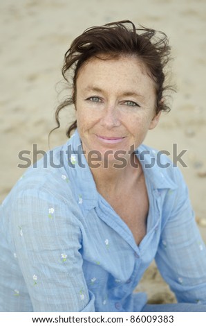 A happy smiling woman in her forties sitting on the beach wearing a blue blouse.