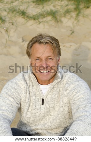 A color portrait of a happy smiling blonde haired man in his forties sitting on the beach.