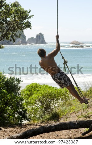 A man standing on a rope swing and admiring the beautiful sea view during his summer vacation.