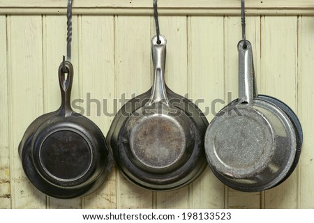 Old iron skillets and antique pans hang on iron hooks against a wall.