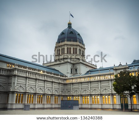The Australian flag flies over the central dome of the historic Melbourne landmark Royal Exhibition Building. The building\'s interior lighting glows golden through the gloom of a wet Autumn morning.