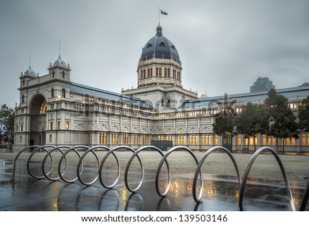 A bicycle parking spiral outside the historic Melbourne landmark Royal Exhibition Building. The building's interior lighting glows golden through the gloom of a wet Autumn morning.
