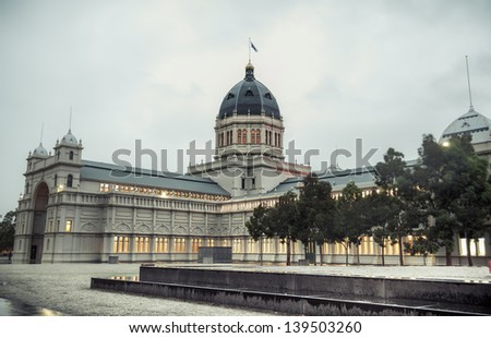 The Australian flag flies over the central dome of the historic Melbourne landmark Royal Exhibition Building. The building\'s interior lighting glows golden through the gloom of a wet Autumn morning.