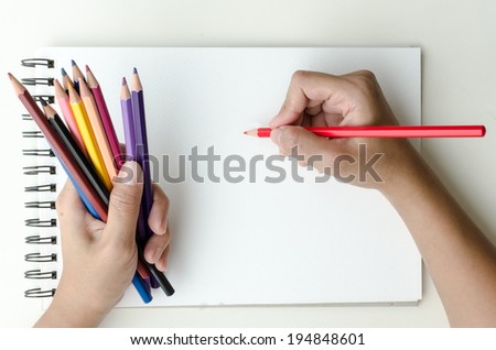 Man holding a fistful of colored pencils in one hand while commencing sketching in a sketch book to show off his creativity and artistic skills with the other, view from above