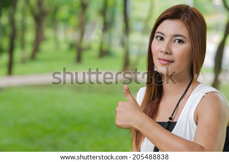 Attractive young Asian woman giving a thumbs up gesture of approval and success as she stands outdoors in a park