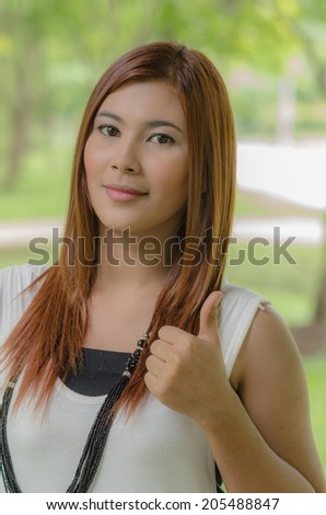 Attractive young Asian woman giving a thumbs up gesture of approval and success as she stands outdoors in a park