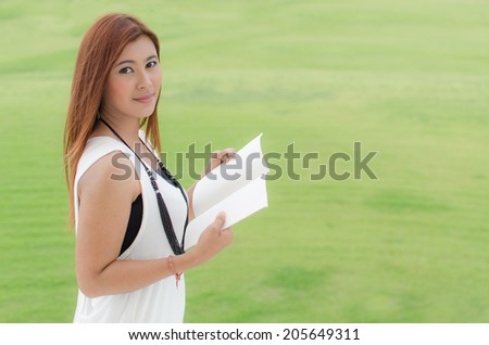 Beautiful young redhead Asian woman holding a book while standing outdoors in a park looking at camera