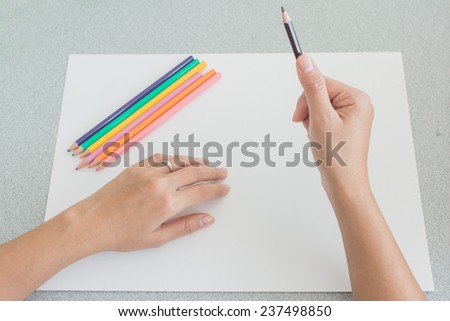 Artist preparing to sketch with colored pencil crayons, close up high angle view of his hands holding a pencil with the rest of the set lying on a large blank white sheet of art paper