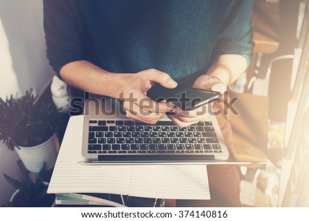 Man holding a smartphone in hands and writes the message. Generic design laptop is on his knees. Sunlights