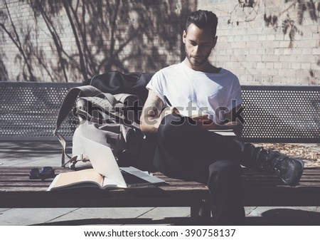 Photo bearded artist wearing white tshirt sitting city park and draws. Studying at the University, working project. Books, generic design laptop, backpack bench.  Horizontal