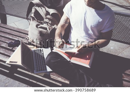 Closeup photo man wearing white tshirt sitting city park and writing notebook. Studying at the University,  preparation for exams. Books, generic design laptop, backpack bench. Horizontal, film effect