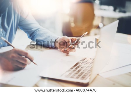 Working process photo. Manager working wood table with new  project. Modern notebook table. Pencil holding hand, typing  contemporary smartphone. Horizontal. Flares, film effect. Blurred background